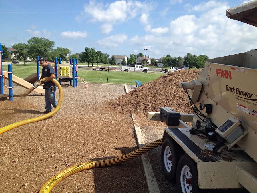 bark and mulch blowing 101: A person holds a yellow hose and dispenses bark mulch on a playground, using a Finn Bark Blower.