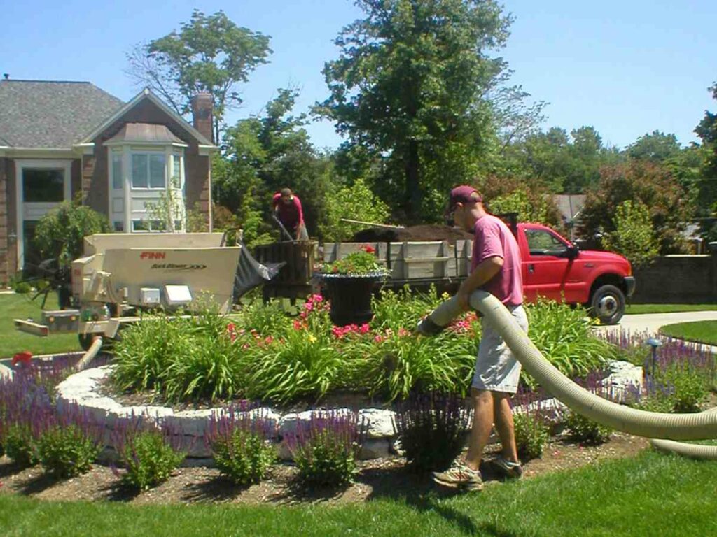 A person holding a thick hose blows mulch onto a circular garden feature. A Finn mulch blower sits in the background.