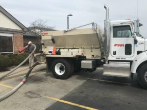 Bark & Mulch Blower Machine Safety for Landscaping Crews. A Finn material blower is sits in a parking lot. A person is holding the blower hose over their shoulder and blowing mulch around a bush.