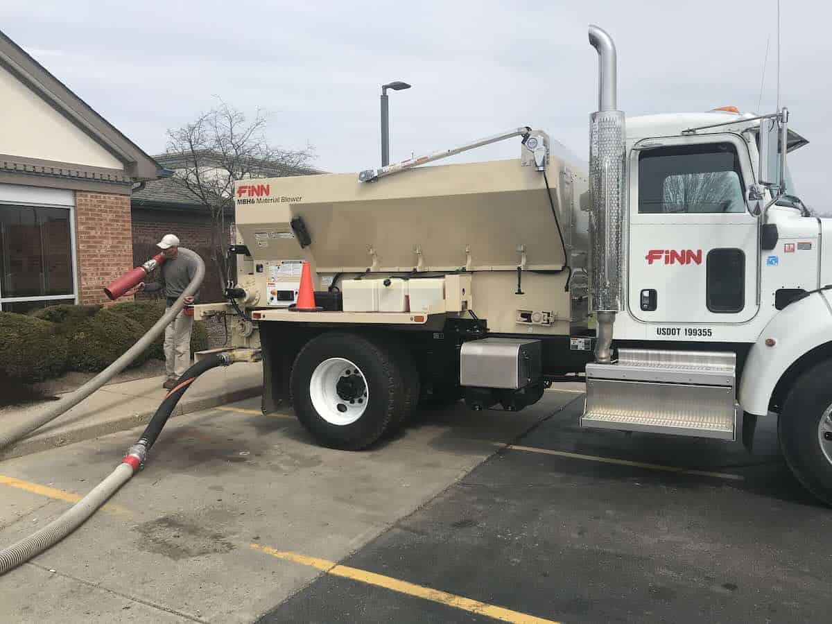 Bark & Mulch Blower Machine Safety for Landscaping Crews. A Finn material blower is sits in a parking lot. A person is holding the blower hose over their shoulder and blowing mulch around a bush.