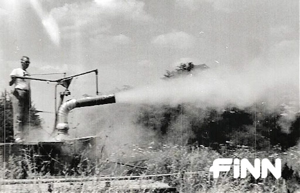 A black and white photo depicts straw blown across a field out of a spout. A person stands on top of a platform watching.