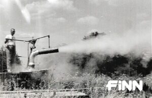 A black and white photo depicts straw blown across a field out of a spout. A person stands on top of a platform watching.